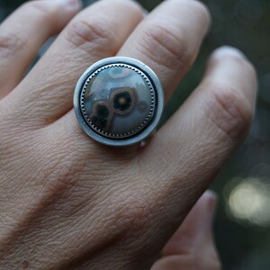 May include: A large, round ring with a silver band. The ring features a central stone with a unique pattern of brown, green, and cream colours. The stone is set within a textured silver bezel.