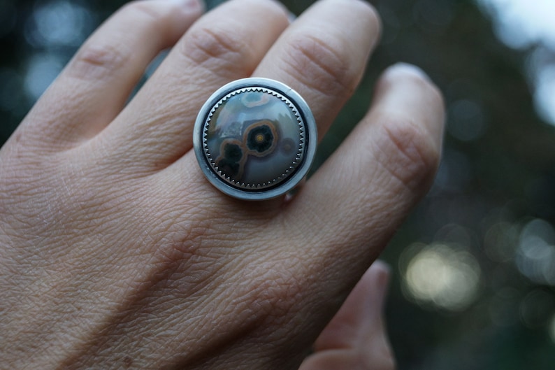 May include: A large, round ring with a silver band. The ring features a central stone with a unique pattern of brown, green, and cream colours. The stone is set within a textured silver bezel.