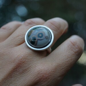 May include: A silver ring featuring a large, circular stone with a distinctive pattern. The stone displays grey, brown, and blue hues in a concentric design. The ring has a plain silver band and setting, highlighting the stone's unique appearance.