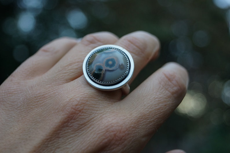 May include: A silver ring featuring a large, circular stone with a distinctive pattern. The stone displays grey, brown, and blue hues in a concentric design. The ring has a plain silver band and setting, highlighting the stone's unique appearance.