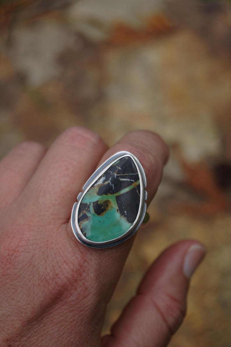 May include: A silver ring featuring a large, teardrop-shaped stone. The stone has a striking pattern of black, white, and green hues. The ring's setting is a simple, polished silver, highlighting the natural beauty of the stone.