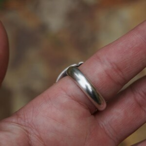 May include: A silver ring with a rounded band and a decorative top, worn on a finger. The ring has a polished, reflective surface, and the design on top is slightly visible. The background is blurred, suggesting an outdoor setting.