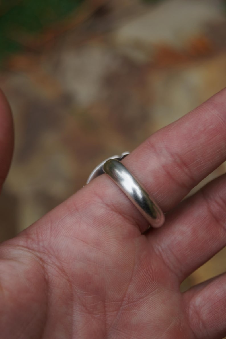 May include: A silver ring with a rounded band and a decorative top, worn on a finger. The ring has a polished, reflective surface, and the design on top is slightly visible. The background is blurred, suggesting an outdoor setting.