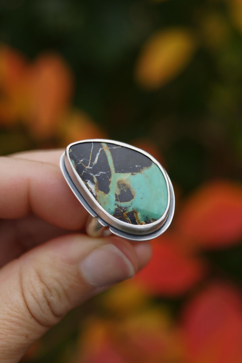 May include: A silver ring featuring a large, teardrop-shaped stone. The stone has a striking pattern of black, turquoise, and brown hues. The ring's setting is a simple, polished silver band, highlighting the unique stone.