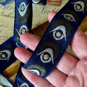 May include: Blue and green patterned ribbon with a repeating design of stylized eyes. The ribbon is woven with a textured surface.