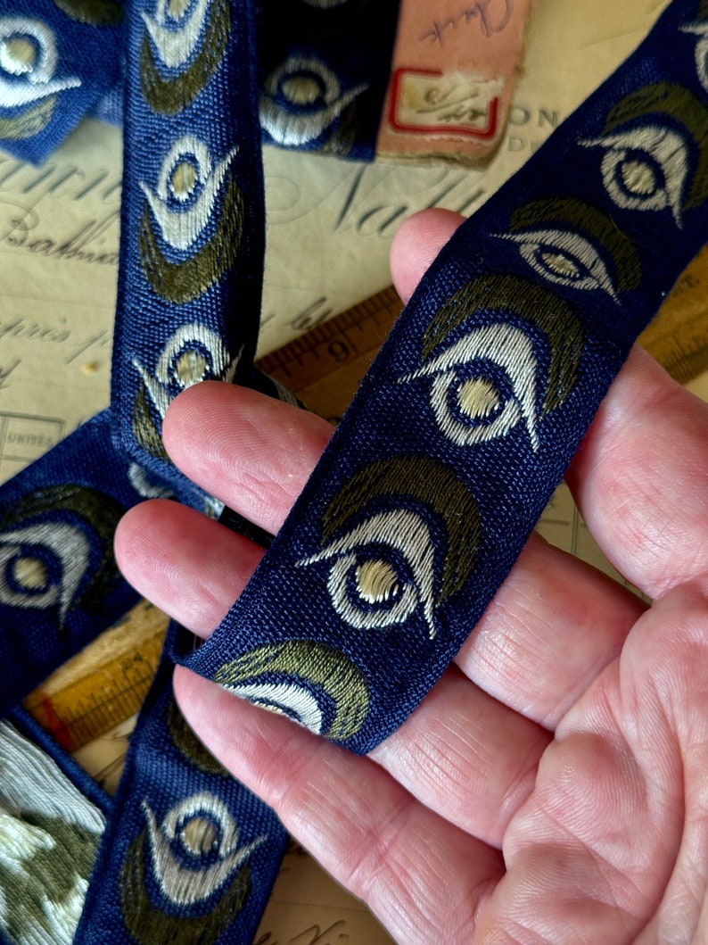May include: Blue and green patterned ribbon with a repeating design of stylized eyes. The ribbon is woven with a textured surface.