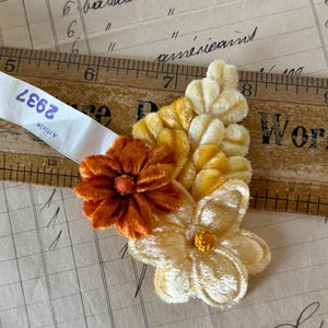 May include: A fabric floral applique featuring a rust-orange velvet flower, a pale yellow velvet flower, and a cluster of yellow velvet leaves. A wooden ruler is positioned behind the applique, showing a width of approximately 2.5 inches.