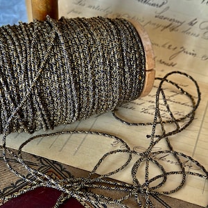 Puede incluir: Un carrete de cordón negro y dorado con un centro de madera. El cordón está enrollado firmemente alrededor del carrete y está listo para ser utilizado para manualidades o decoración.