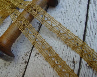 May include: Close-up of two strands of gold-colored decorative trim. The trim has a delicate, open weave design with repeating geometric shapes. The trim is lying on a wooden spool, suggesting it is used for crafts or embellishments.