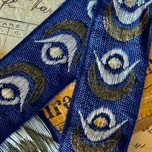 May include: Blue fabric ribbon with a repeating pattern of white and gold peacock eyes. The fabric has a textured woven look.