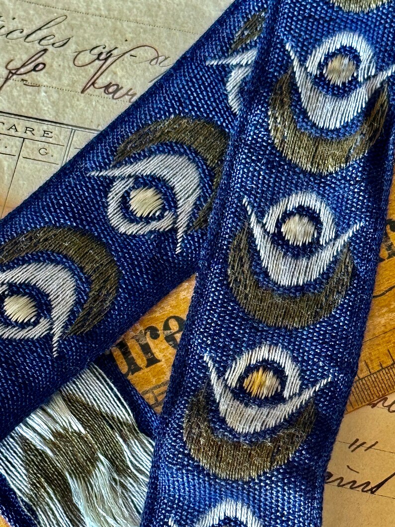 May include: Blue fabric ribbon with a repeating pattern of white and gold peacock eyes. The fabric has a textured woven look.