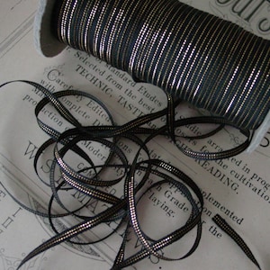 May include: A spool of black and gold ribbon with a silver metallic thread. The ribbon is wound around a spool and is lying on a piece of paper with text.