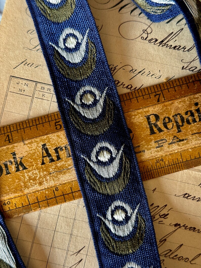 May include: Blue and green patterned ribbon with white accents. The ribbon has a repeating design of stylized eyes. The ribbon is lying on top of a wooden ruler with the words "York Arm" and "Repair" printed on it.