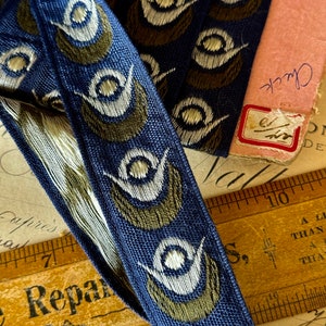May include: Blue and gold ribbon trim with a repeating pattern of stylized white and gold eyes. The ribbon is lying on a wooden ruler and a piece of paper with writing.