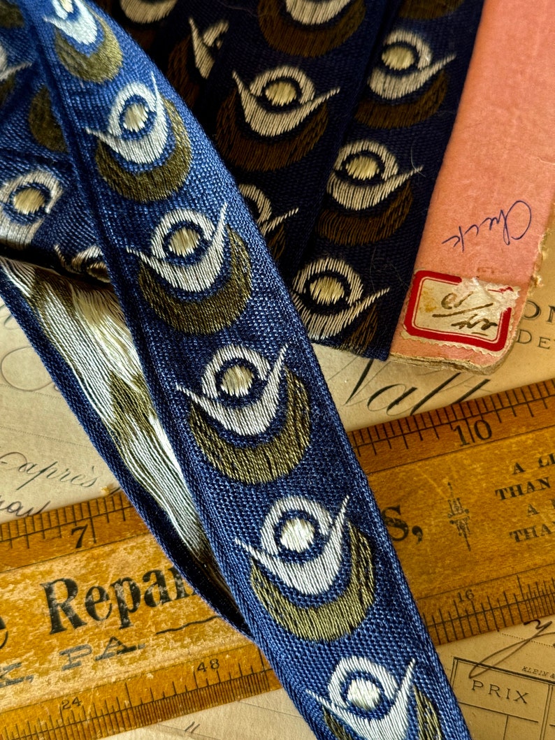 May include: Blue and gold ribbon trim with a repeating pattern of stylized white and gold eyes. The ribbon is lying on a wooden ruler and a piece of paper with writing.