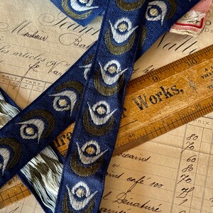 May include: Two blue and white ribbon trims with a peacock eye design. The ribbon is woven with a dark blue background and white and green peacock eyes. The ribbon is lying on top of a vintage ledger with handwritten text.