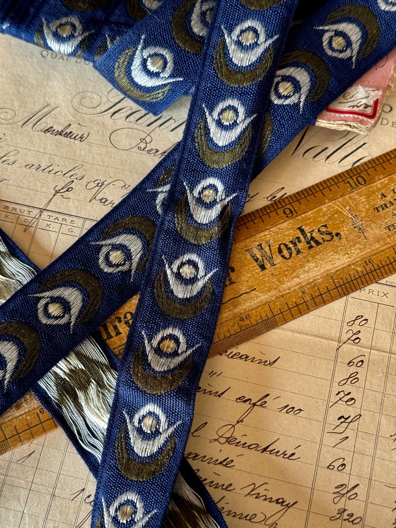 May include: Two blue and white ribbon trims with a peacock eye design. The ribbon is woven with a dark blue background and white and green peacock eyes. The ribbon is lying on top of a vintage ledger with handwritten text.