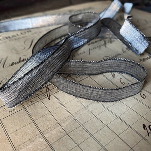 May include: Silver ribbon with a metallic sheen, edged with a darker trim, is arranged in loose loops. The ribbon is lying on a vintage ledger with handwritten text and numbers, creating a contrast between textures and eras.