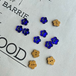 May include: Assortment of blue and gold flower-shaped buttons. The blue buttons are made of glass and have a shiny finish. The gold buttons are made of wood and have a matte finish.
