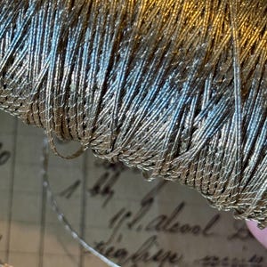 Puede incluir: Primer plano de una bobina de hilo metálico plateado y dorado. El hilo está enrollado apretado y tiene un acabado brillante y reflectante.