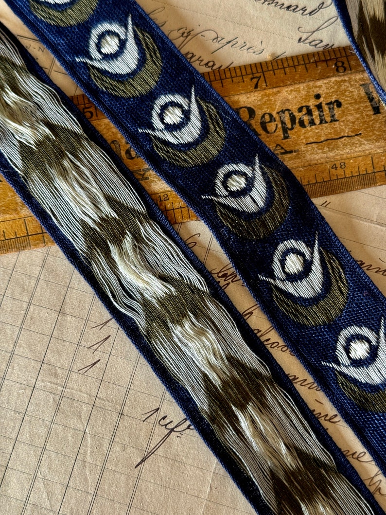 May include: Two blue and white patterned ribbon trims with a repeating design of stylized eyes. The ribbon trims are on a brown surface with a ruler and a piece of paper with the word "Repair" written on it.