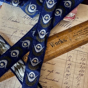 May include: Close-up of blue and green patterned ribbon with white accents. The ribbon has a repeating design of stylized eyes. The ribbon is lying on top of a vintage ledger and a wooden ruler with the words "Works" printed on it.