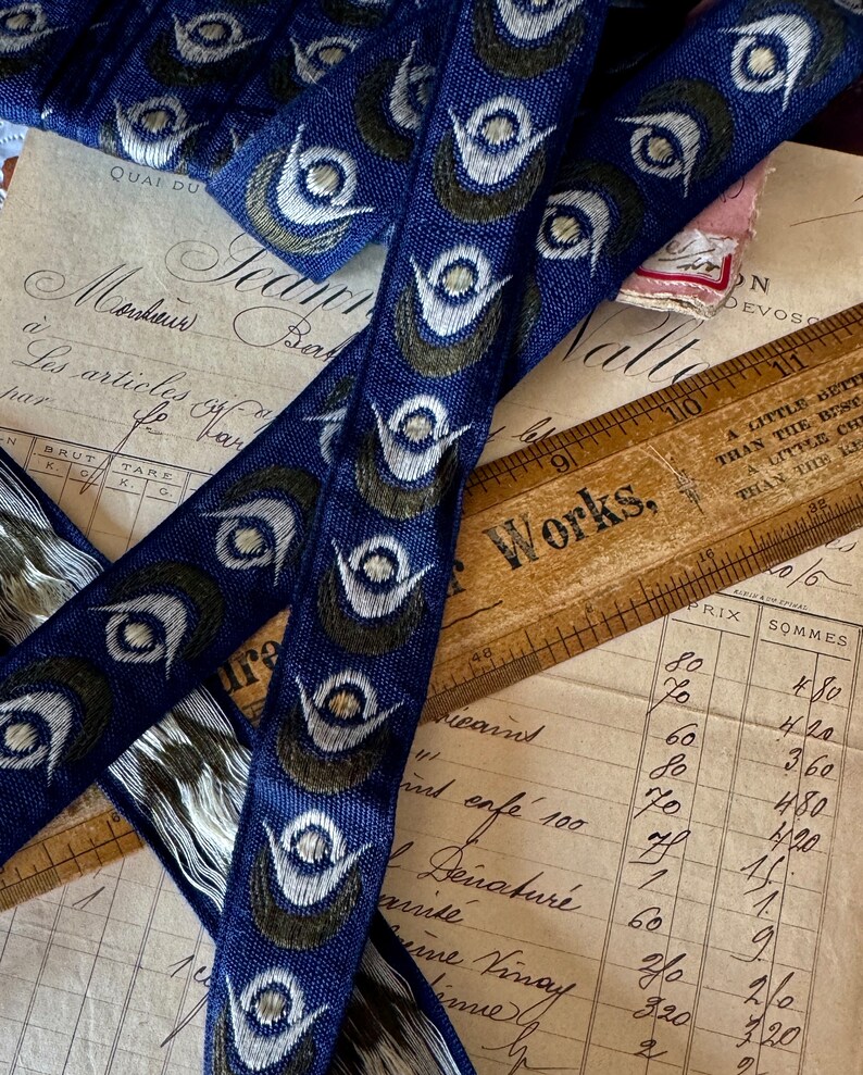 May include: Close-up of blue and green patterned ribbon with white accents. The ribbon has a repeating design of stylized eyes. The ribbon is lying on top of a vintage ledger and a wooden ruler with the words "Works" printed on it.