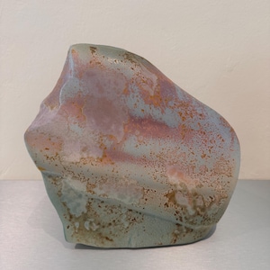 May include: A polished, irregularly shaped stone featuring a smooth surface with a blend of muted sage green, soft lavender, and warm ochre-coloured mineral inclusions that create a mottled, organic pattern across the entire face of the rock.