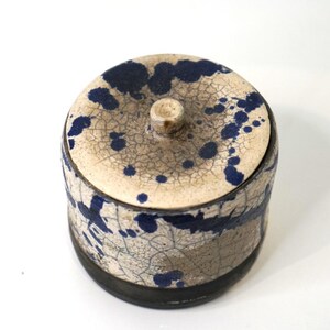 May include: A cylindrical ceramic jar with a lid, featuring a cream-colored base with a dark blue speckled pattern and a fine crackle glaze finish. The base of the jar is a solid dark brown, and the lid includes a small, centered circular knob.