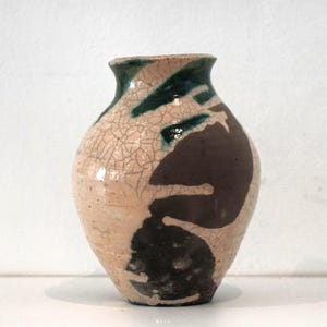 Puede incluir: Un jarrón de cerámica hecho a mano con un esmalte craquelado único. El jarrón presenta una base rosa claro con diseños abstractos marrón oscuro y verde. Tiene un cuerpo redondeado y un cuello estrecho, perfecto para exhibir flores o como pieza decorativa.