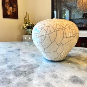May include: A rounded ceramic vase with a cream-colored glaze featuring a prominent dark gray crackle pattern. The vessel has a wide, bulbous body that tapers toward a narrow base and a small, circular opening at the top, showcasing a distinct texture.