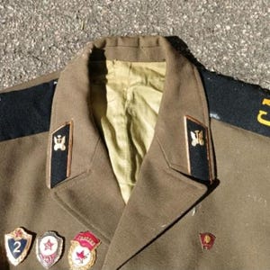 May include: An olive drab military jacket featuring black shoulder boards with the yellow Cyrillic letters CA, black collar tabs with gold emblems, and several metal badges pinned to the chest. A patch is visible on the upper left sleeve.