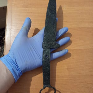 Puede incluir: Una daga negra de estilo antiguo, con una hoja puntiaguda y un mango con un lazo en el extremo. La daga tiene una superficie texturizada y parece ser de metal. El diseño evoca un tema histórico o medieval.