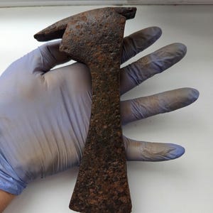 Puede incluir: Un hacha de metal oxidado y envejecido, con diseño de cabeza de martillo. El filo del hacha es rectangular con un borde puntiagudo y un martillo en el lado opuesto. El hacha es sostenida por una mano enguantada, mostrando su aspecto desgastado.