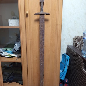 Puede incluir: Una espada larga y antigua con una hoja y guarda oxidadas de color marrón. La empuñadura presenta un pomo circular. La espada se exhibe verticalmente contra un armario de madera. El aspecto general sugiere un tema histórico o medieval.