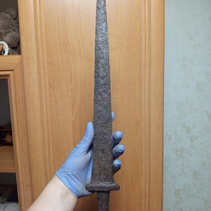May include: A long, antique sword with a pointed tip and a textured, dark brown surface. The sword has a crossguard and a handle with a rounded base. The blade appears to be made of metal and shows signs of age and wear.