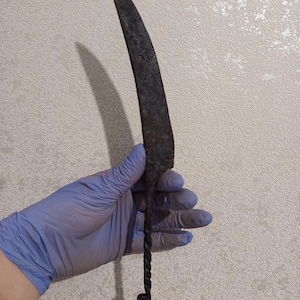 Puede incluir: Un cuchillo de metal oxidado y curvado, con un mango retorcido y un pequeño lazo en el extremo. La hoja es oscura y parece envejecida. El cuchillo lo sostiene una persona con un guante azul.