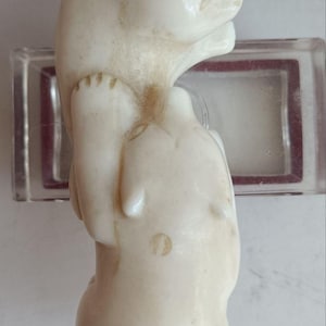 May include: A smooth, off-white carved stone figurine depicting two stacked animal figures. The upper figure has a rounded head and snout, while the lower figure is bulbous and rounded. The surface shows subtle natural variations and light brown markings.