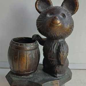 Può includere: Una statuetta in metallo color bronzo raffigurante un orso in piedi accanto a un piccolo barile. L'orso ha orecchie arrotondate e una superficie ruvida, mentre il barile presenta doghe verticali. L'intero pezzo è montato su una base solida.