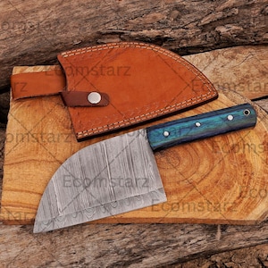 May include: A Damascus steel cleaver with a blue and green handle rests on a wooden cutting board. A brown leather sheath with a snap closure is positioned above the cleaver. The cleaver blade has a curved edge.