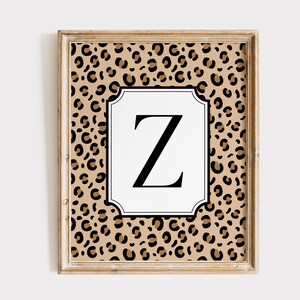 May include: A framed wall art print featuring a tan and black leopard print background with a central white rectangular plaque with scalloped edges. The letter Z is printed in a bold black serif font in the center of the white plaque.
