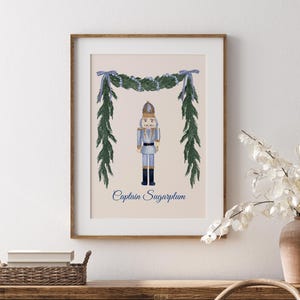 May include: A framed print featuring a watercolor illustration of a nutcracker soldier, with the text "Captain Sugarplum" below. The artwork is decorated with a garland and bows. The print is displayed on a wooden dresser.