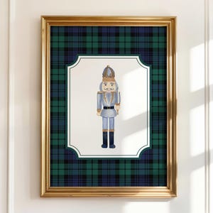 May include: A framed print featuring a watercolor illustration of a nutcracker soldier. The artwork is set against a green and blue plaid background within a gold frame. The nutcracker is depicted in shades of blue, gold, and black.