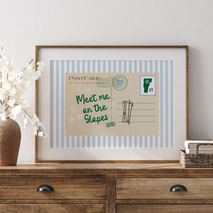 May include: A framed postcard print with a brown wooden frame. The postcard has a light blue and white striped background. The text on the postcard reads "Meet me on the Slopes" with snowflakes and ski graphics. A Vermont stamp is visible.