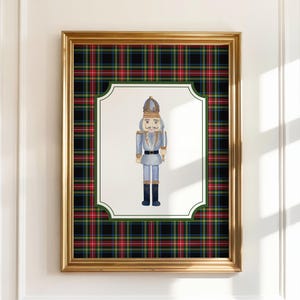 May include: A framed print featuring a watercolor illustration of a nutcracker soldier. The artwork is set against a white background with a green border, framed by a gold frame and a plaid pattern in red, green, and black.