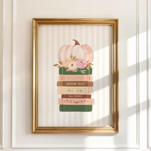 May include: A framed print featuring a stack of books topped with a light pink pumpkin and floral arrangement. The books are in shades of green, brown, and pink, with the words "AUTUMN TALES" and "FALL STORIES" visible. The frame is gold.