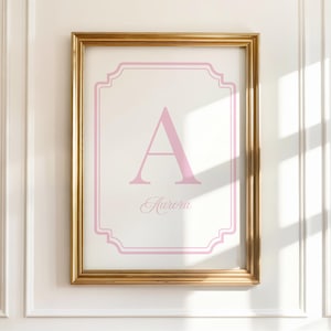 May include: A framed print featuring a pink letter "A" and the name "Aurora" in a delicate script. The artwork is set against a white background within a pink decorative border, encased in a gold frame. The framed print is mounted on a white wall.