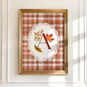 May include: Framed wall art featuring a decorative letter "A" in a burgundy color, surrounded by autumn leaves. The letter is set against a white oval with a lace-like border, on a plaid background in shades of brown and white. The frame is gold.