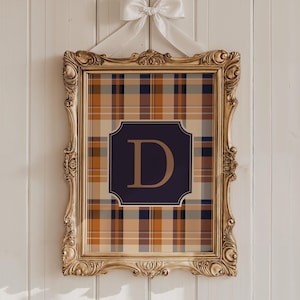 May include: A framed wall art piece featuring a plaid pattern in shades of tan, navy, and orange. The artwork includes a navy blue square with the letter "D" in a gold color. The frame is gold and ornate, with a white bow at the top.