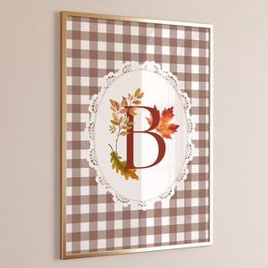 Puede incluir: Arte de pared enmarcado con un patrón de cuadros marrones y blancos. Un diseño ovalado con un borde de encaje contiene la letra "B" en rojo oscuro, con hojas y follaje otoñal.